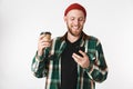 Portrait of european guy wearing plaid shirt holding paper cup with coffee and using mobile phone, while standing isolated over Royalty Free Stock Photo