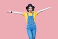 Portrait of excited beautiful young woman in yellow t-shirt, blue denim overalls, makeup, black hat standing with raised arms and