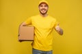 Portrait of excited delivery man in yellow uniform holding paper box isolated over yellow background.