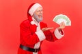 Portrait of excited elderly man with gray beard wearing santa claus costume holding fan of euro in hands, pointing at money, keeps Royalty Free Stock Photo