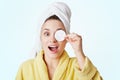 Portrait of excited female with towel on head, covers eye with cotton wheel, demonstrates her care about beauty, prepares for date