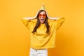 Portrait of excited joyful young woman in orange heart glasses and birthday party hat screaming, putting hands on head