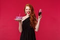 Portrait of excited laughing gorgeous redhead woman having fun at b-day party, holding glass of wine and birthday cake