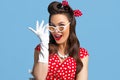 Portrait of excited pin up woman in polka dot dress, headband and gloves touching her sunglasses on blue background Royalty Free Stock Photo