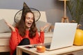 Portrait of excited positive woman wearing witch hat sitting in front on laptop in home interior and having video call or