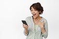 Portrait of excited and surprised young woman looking at mobile phone happy, laughing amazed at message, standing Royalty Free Stock Photo