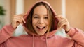 Portrait of excited tenage girl wearing hood and smiling at camera, posing indoors at home interior