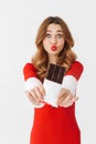 Portrait of excited woman 20s wearing Santa Claus red costume smiling and eating chocolate bar, isolated over white background Royalty Free Stock Photo