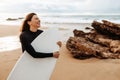 Portrait of excited young surfer woman on the beach holding her surfboard, posing and smiling, enjoying water sport