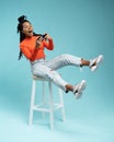 Portrait of an excited young woman sitting on a stool and playing games on mobile phone  over blue background. Royalty Free Stock Photo