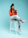 Portrait of an excited young woman sitting on a stool and playing games on mobile phone  over blue background. Royalty Free Stock Photo