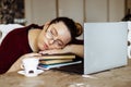 Portrait of exhausted middle-aged woman sitting at table near cup, laptop, sleeping on crossed arms on pile of books.