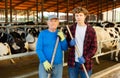 Elderly farmer standing with teenage grandson near stall with cows