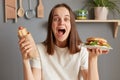 Portrait of extremely happy amazed woman with dark hair dressed in white T-shirt holding big burger sandwich and sausage in dough Royalty Free Stock Photo