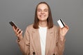 Portrait of extremely happy woman with brown hair wearing beige jacket, using cell phone and showing credit card, online shopping Royalty Free Stock Photo