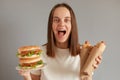 Portrait of extremely happy woman eating fast food, holding hot dog and sandwich wearing white T-shirt posing isolated over gray Royalty Free Stock Photo