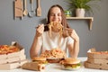 portrait of extremely hungry cheerful Caucasian woman wearing white casual T-shirt sitting at table in kitchen, being happy to eat Royalty Free Stock Photo