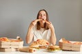 Portrait of extremely hungry woman with brown hair wearing white T-shirt sitting at table over gray background, biting