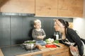 Portrait of family of little boy, young woman cooking soup in kitchen. Baby toddler sitting on table near black cooker. Royalty Free Stock Photo