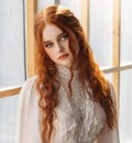 Portrait fantasy beauty red-haired woman green eyes looking at camera. White old style vintage dress. Stads by window Royalty Free Stock Photo