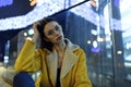 Portrait of fashion female model posing in yellow jacket and glasses, in city lights by night. Womenswear style