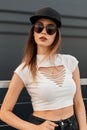 Portrait fashion pretty urban woman in white stylish torn top in jeans in cool trendy sunglasses and trendy baseball black cap on Royalty Free Stock Photo