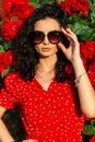 Portrait of fashionable pretty woman with sunglasses and stylish silver bracelet in elegant red polka dot dress posing near bush Royalty Free Stock Photo