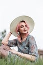 Portrait fashionable pretty young woman model with chic pink hair in hat in stylish blue dress with floral print on grass in field Royalty Free Stock Photo