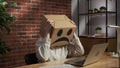 Portrait of female in cardboard box with emoji on head. Worker sitting at the desk working on laptop, frustrated