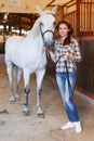 Female farmer standing with white horse at stabling indoor Royalty Free Stock Photo