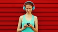 Portrait fitness smiling young woman in headphones listening to music with smartphone on red background