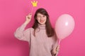 Portrait of flirty attractive winking dark haired woman with pink balloon, dresses warm rose sweater, posing against rosy studio