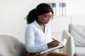 Portrait of focused black female psychologist writing in clipboard, sitting in armchair at modern office Royalty Free Stock Photo
