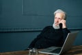 Portrait of focused gray-haired senior aged businessman talking on mobile phone sitting at wooden table with laptop Royalty Free Stock Photo