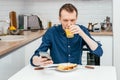 Portrait of focused middle-aged man in blue shirt looking at mobile phone drinking orange juice in white modern kitchen. Royalty Free Stock Photo