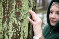 Portrait of focused middle-aged woman touching green moss on trunk of tree with hand in park forest in snowy winter. Royalty Free Stock Photo