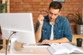 Portrait of focused thinking young man reading paper book holding end of glasses near mouth sitting at desk with desktop Royalty Free Stock Photo