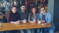 Portrait of four young people friends in casual clothes sitting at table in spacious cafe with tea cups and looking at