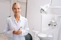 Portrait of friendly blonde female dental orthodontist doctor standing near dental chair in office holding tools for Royalty Free Stock Photo