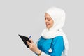 Friendly, Muslim doctor or nurse woman in a hijab with a stethoscope writes a prescription on pills on a gray background