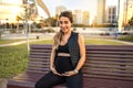 Portrait of friendly young future mother holding and touching her stomach while resting on bench outdoors. Royalty Free Stock Photo