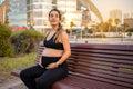 Portrait of friendly young pregnant woman touching her stomach while resting on bench outdoors. Royalty Free Stock Photo