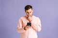 Portrait of frustrated young man holding phone with doubtful and skeptical expression on pink  background. Royalty Free Stock Photo