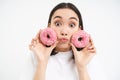 Portrait of funny asian woman, tempted to eat tasty glazed dougnut, holding two donnuts and smiling excited, isolated on