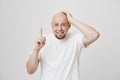 Portrait of funny bald european male with beard holding hand on head while pointing up with another, smiling and Royalty Free Stock Photo