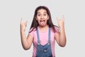 Portrait of funny beautiful brunette young girl in casual pink t-shirt and blue overalls with rock and roll hand horns and tongue