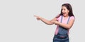 Portrait of funny beautiful brunette young girl in casual pink t-shirt and blue overalls standing, pointing at wall empty place Royalty Free Stock Photo