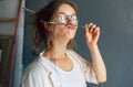 Portrait of a funny beautiful female artist in eyeglasses holding on her lips a painting brush in her workshop. Pretty young woman Royalty Free Stock Photo