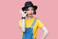 Portrait of funny beautiful young woman in yellow t-shirt and blue denim overalls with makeup and black hat standing with ok sign