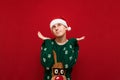 Portrait of a funny bewildered boy in Christmas clothes on a red background, looks up and spreads his hands to the sides. Cute Royalty Free Stock Photo